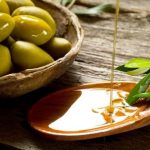 olivový olej na drevenej lyžičke, listy olivovníka a čerstvé olivy v miske